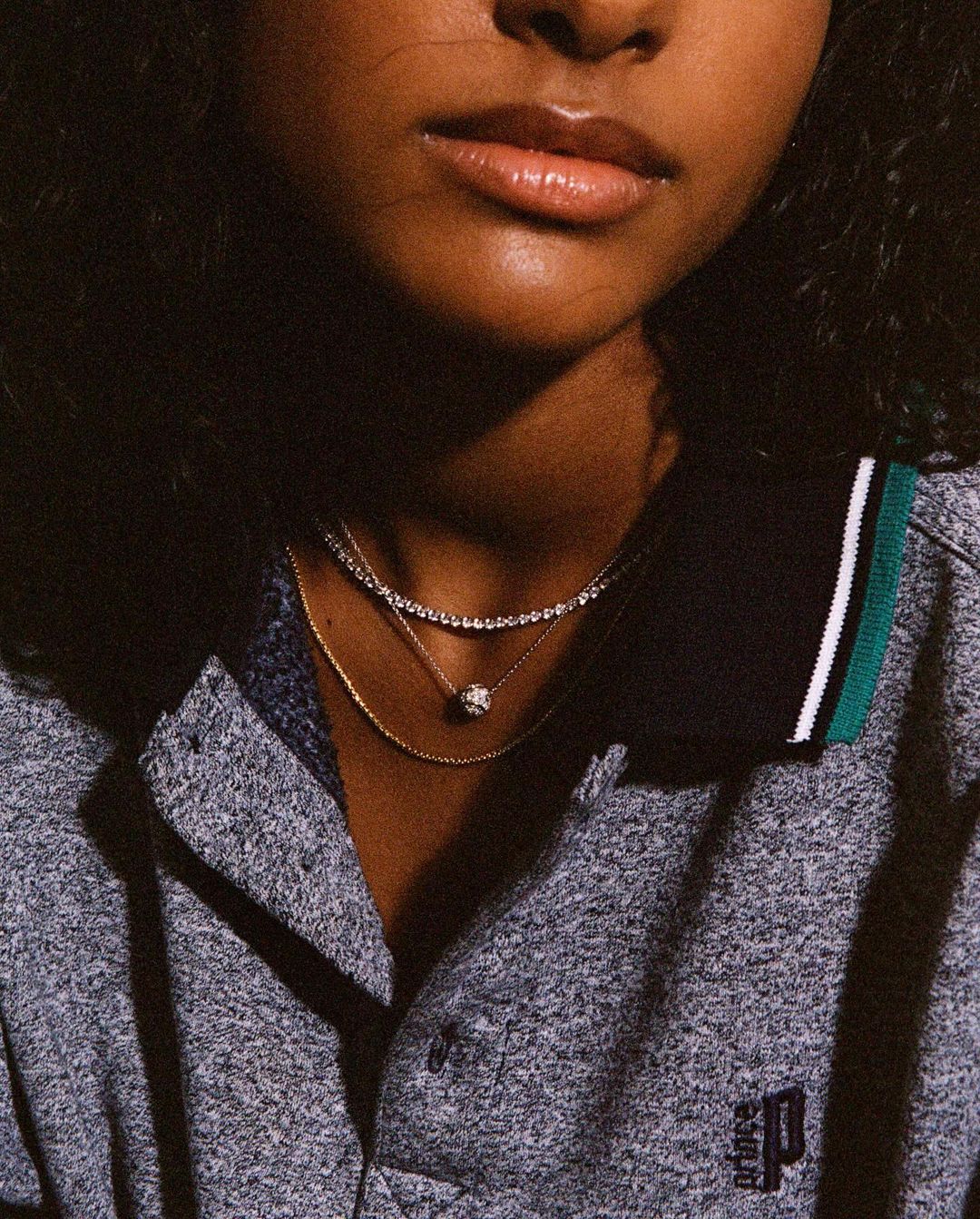 Close-up shot of a young adult woman with layered necklaces wearing a gray polo shirt with dark hair framing her face and shoulders.