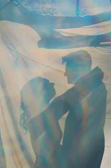 Silhouetted couple embracing under sheer fabric in an ethereal outdoor setting with soft lighting, creating a romantic and intimate portrait.