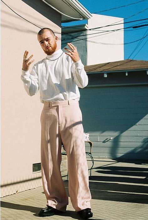Man in white turtleneck and pink pants poses in front of a beige wall outside on a sunny day, hands up, against a residential background.