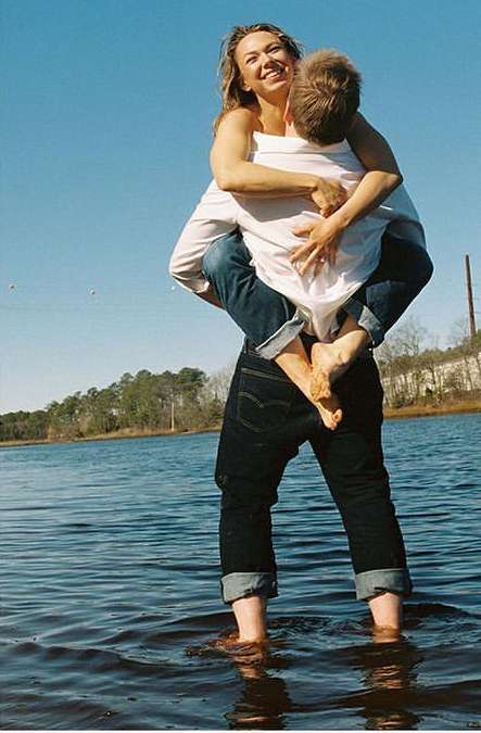 A cheerful woman embraces a man, who carries her in the water of a shallow lake under a sunny sky, with a tree-lined shore in the background.