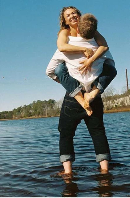 A cheerful woman embraces a man, who carries her in the water of a shallow lake under a sunny sky, with a tree-lined shore in the background.