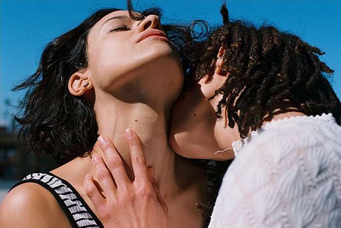 Two adult women pose in close-up on a sunny day. One woman is fair-skinned with dark hair, and the other has dark skin with dreadlocks.