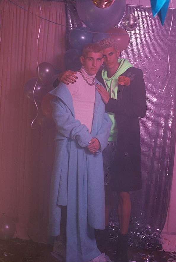 Two young men in formalwear pose in front of a decorated backdrop, illuminated by pink lighting, creating a festive and celebratory ambiance.