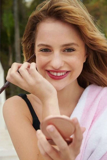 Smiling woman applies eye makeup while holding a compact mirror outdoors with a towel draped over her shoulder in medium portrait shot.