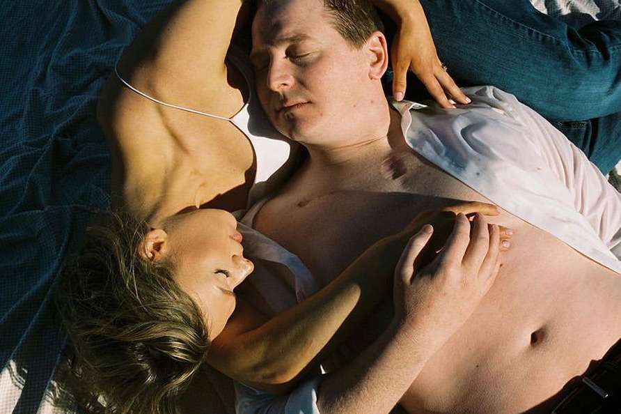 Couple lying down together. Woman with long hair rests her head near a man with eyes closed, wearing an unbuttoned white shirt and jeans.
