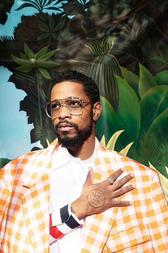 Portrait of an adult man wearing glasses and a checkered blazer, looking off to the side against a backdrop of tropical foliage.
