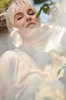Blonde woman with short hair smiles in an outdoor shot, framed by bright light and green foliage. Close-up portrait.