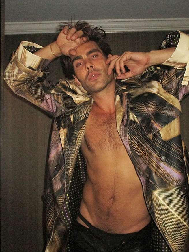 Shirtless man wearing a patterned trench coat with arms raised, photographed by Cully Wright.