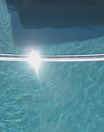 Overhead shot of shimmering water in a turquoise swimming pool with bright sun reflection and a horizontal silver pool rail.