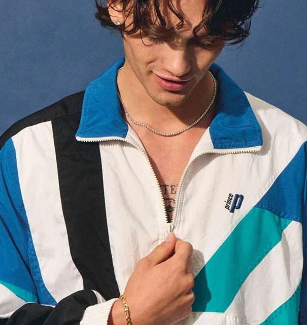 A young adult male poses in a 90s-style windbreaker, with a gold bracelet and thin silver chain. He stands against a blue backdrop.