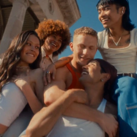 A group of five smiling people huddle together outside against a blue sky and a light-colored building facade in a portrait-style shot.