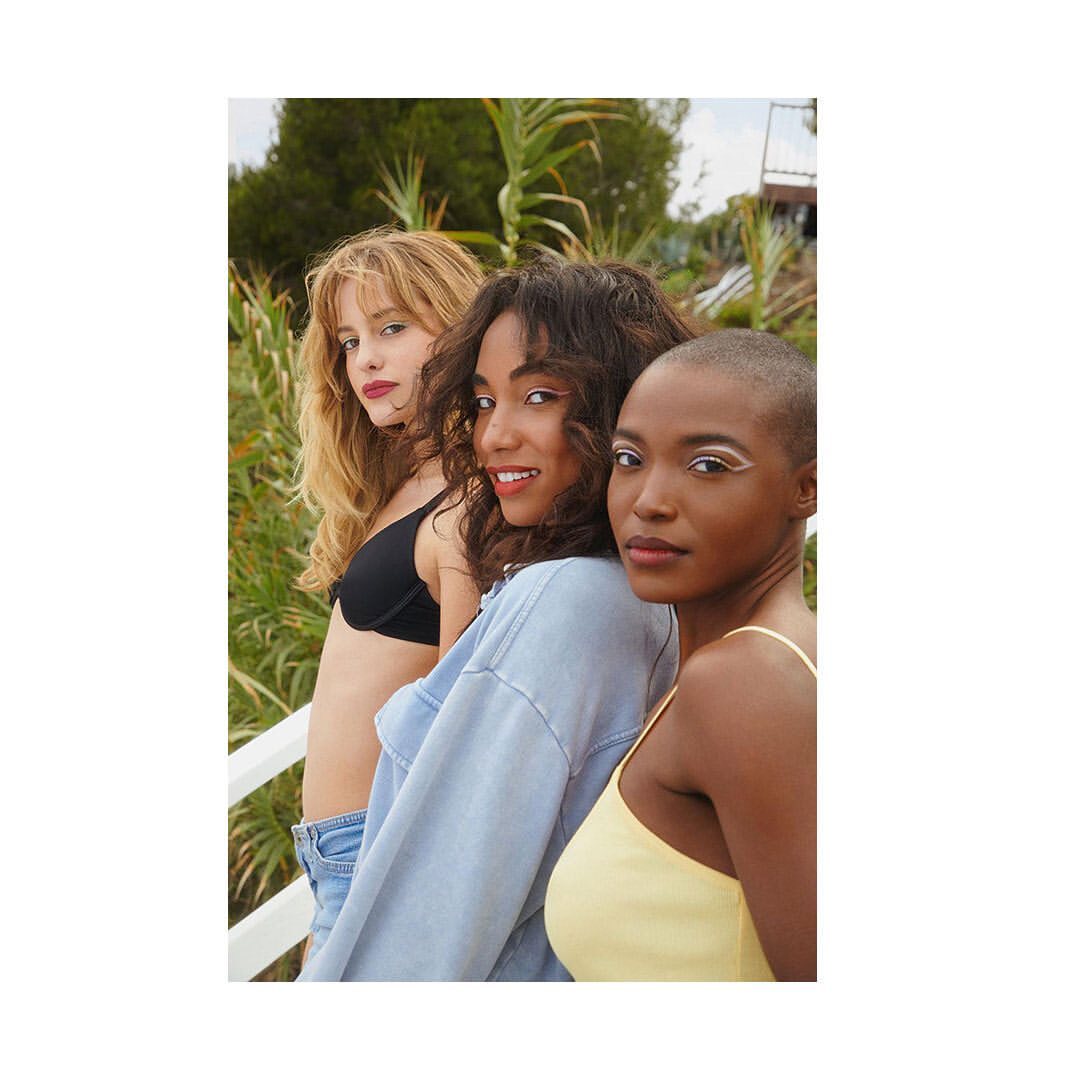 Three female models posing outdoors against a green foliage backdrop in natural daylight.