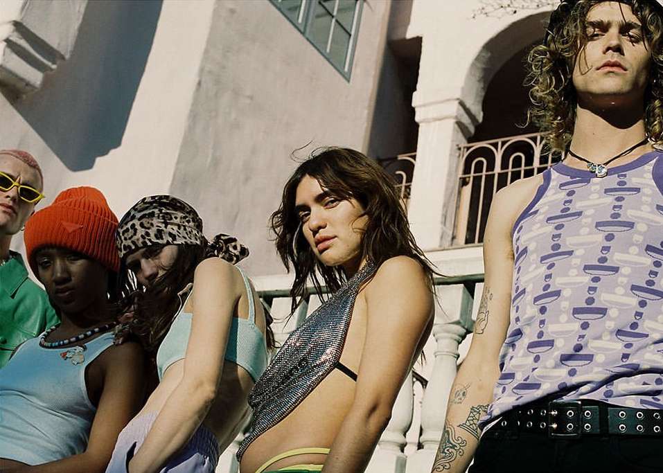 Group of six diverse young adults in urban setting, showcasing fashion styles, posing outdoors against a building with white columns and arches.