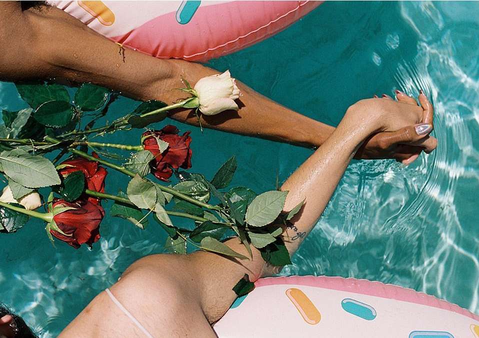 Two individuals, partially submerged in a pool, are holding hands amidst floating roses and colorful donut-shaped inflatables.
