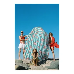Three models posing outdoors against a clear blue sky on a rocky shore, featuring a large striped and floral fabric structure
