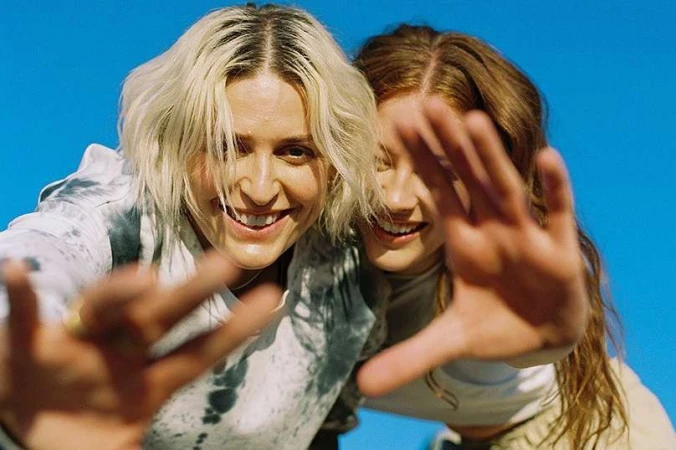 Two smiling women holding their hands out toward the camera against a clear bright blue sky.