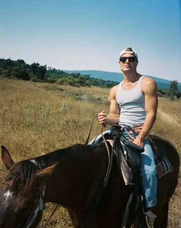 A man wearing sunglasses and a white tank top rides a dark horse through an open grassy field with rolling hills.