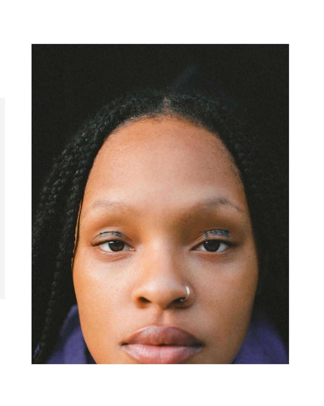 Close-up portrait of a young adult woman with dark braided hair, dark eyes with black eyeliner, a nose ring, and a neutral expression.