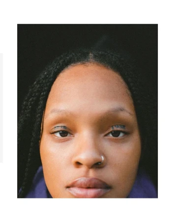 Close-up portrait of a young adult woman with dark braided hair, dark eyes with black eyeliner, a nose ring, and a neutral expression.