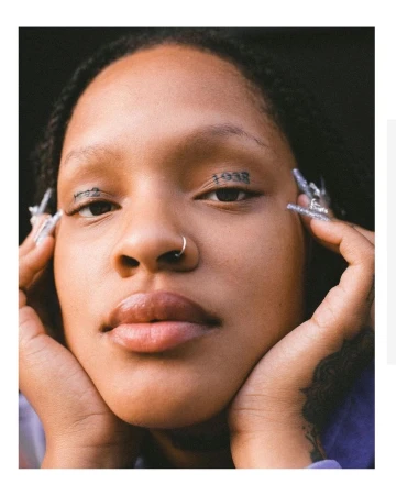 Close up portrait of a Black woman with face tattoos, nose ring, and long glittery nails framing her face.