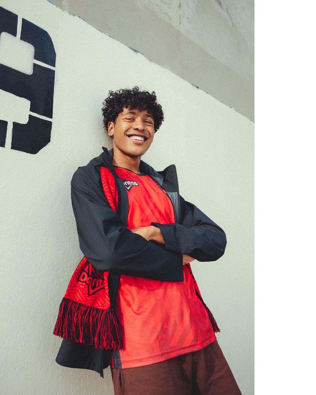 Young man smiling in a red t-shirt and dark jacket posing against a graffiti-covered wall, creating a casual, urban portrait.