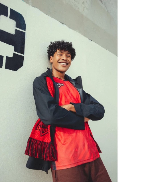Young man smiling in a red t-shirt and dark jacket posing against a graffiti-covered wall, creating a casual, urban portrait.