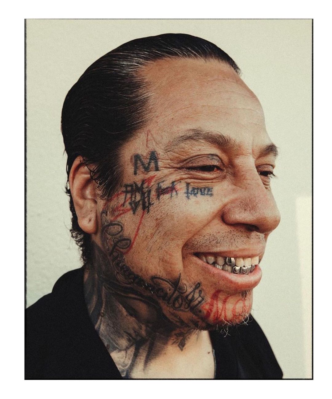 Close-up studio portrait of a smiling adult male with face tattoos, gold teeth, and a black shirt against an off-white background.