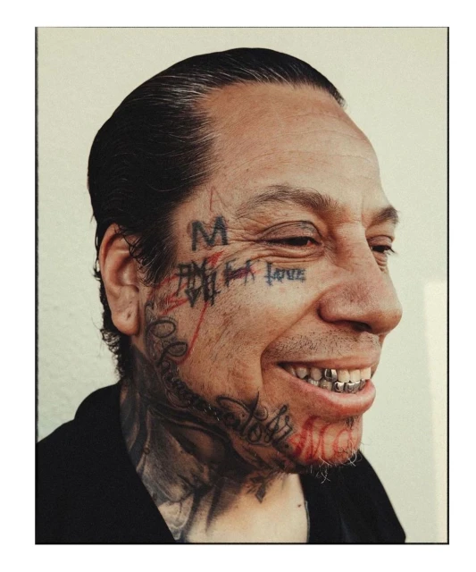 Close-up studio portrait of a smiling adult male with face tattoos, gold teeth, and a black shirt against an off-white background.