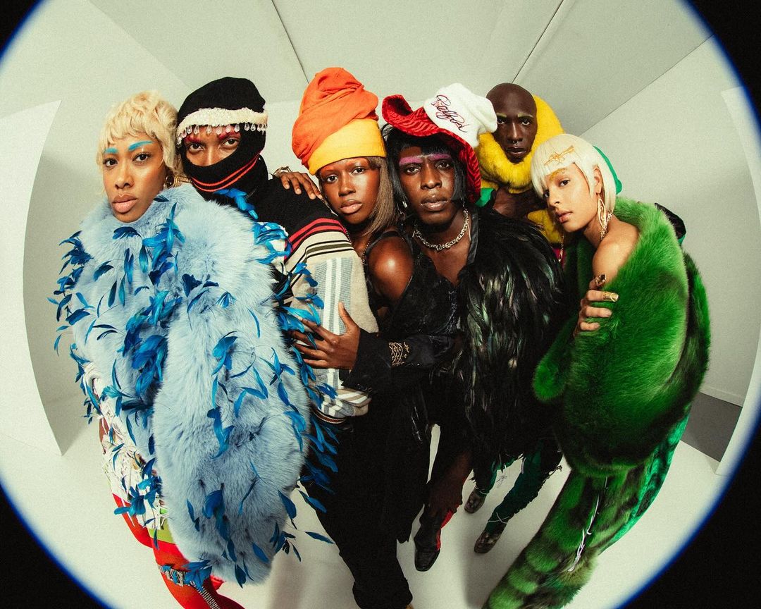 A diverse group of seven adults stand together in a photo studio wearing brightly colored fur coats and unique headwear, captured with a fisheye lens.