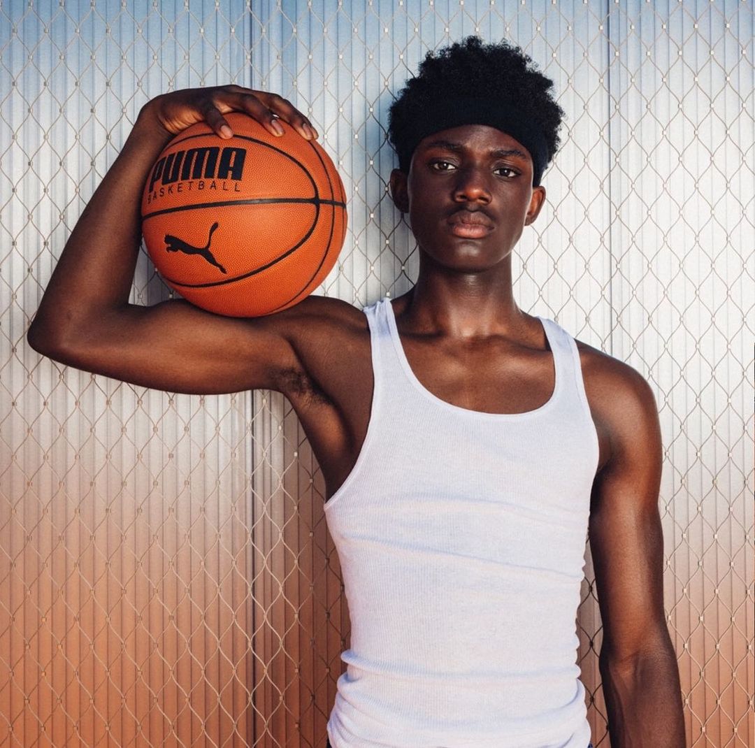 Young Black man in a white tank top holds an orange PUMA basketball in front of a metal-grid background, looking at the camera.