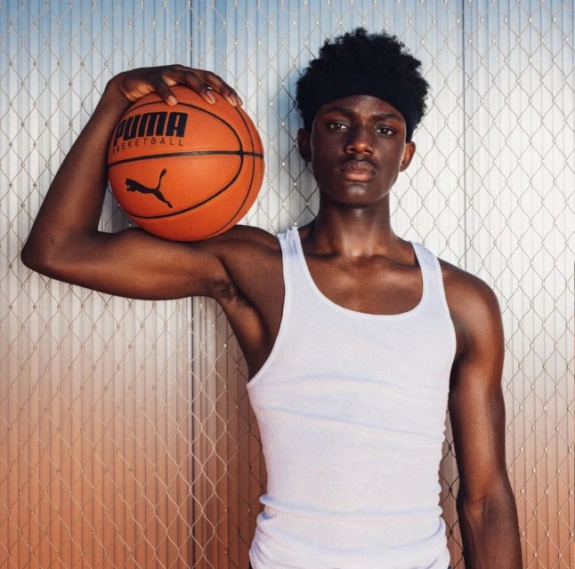 Young Black man in a white tank top holds an orange PUMA basketball in front of a metal-grid background, looking at the camera.