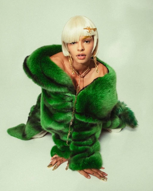 Woman in a vibrant green fur coat and gold jewelry poses in a photo studio, showcasing fashion and beauty against a soft green backdrop.
