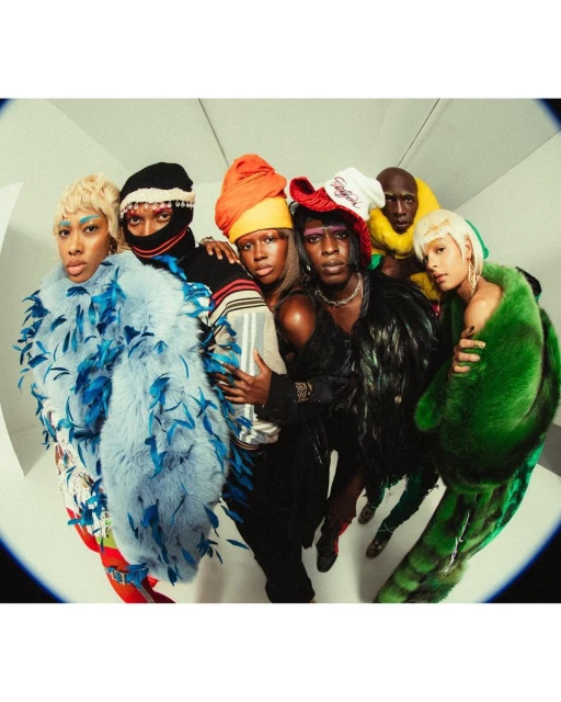 A diverse group of six adults wearing colourful headwear and feather boas, posing together indoors in front of a white wall with a wide fisheye lens.