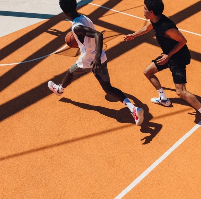 Two young men playing basketball on an orange court with sunlight and shadows. One dribbles a basketball while the other defends.