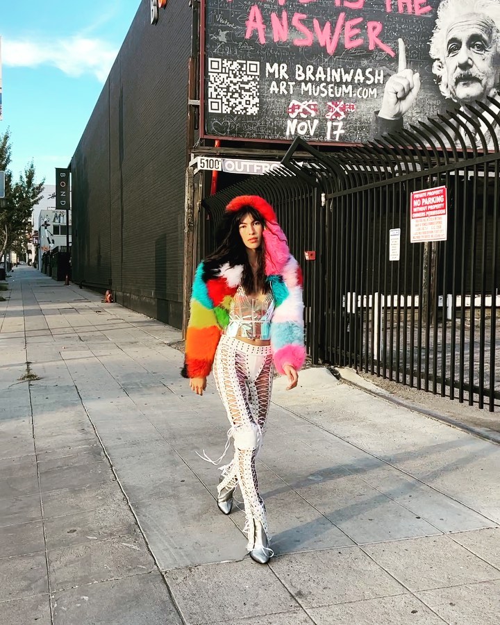 Woman in colorful fur coat and open-weave pants walks along sidewalk in front of street art for Mr. Brainwash Art Museum.