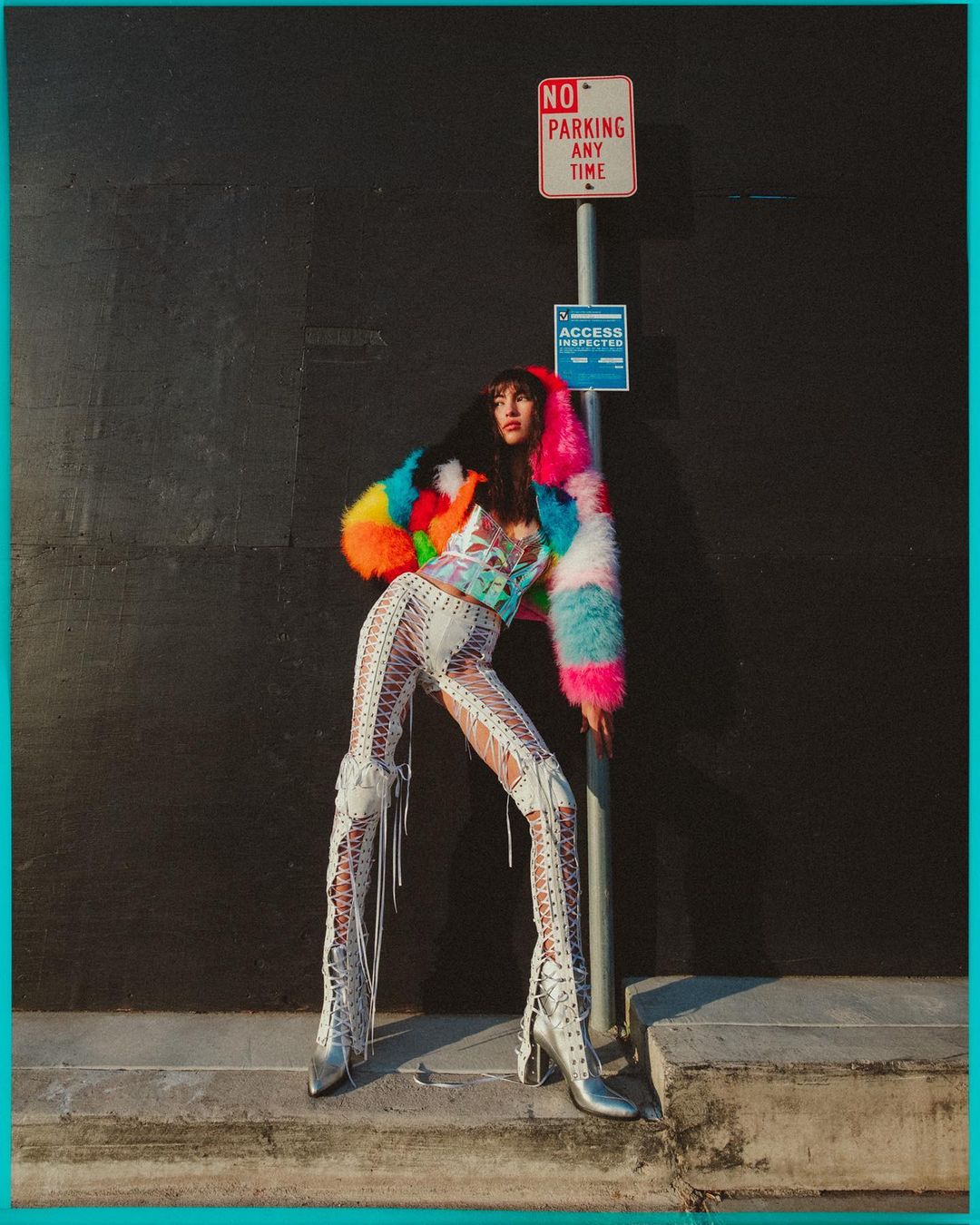 Adult woman in colorful fur coat and lace-up pants poses on sidewalk near a black wall and a parking sign in an urban setting.