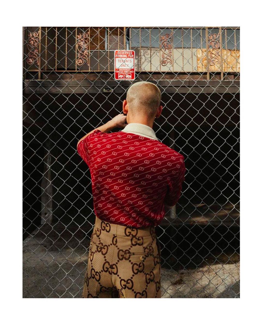 Person with shaved head wearing patterned red shirt and beige patterned pants stands facing away from camera in front of chain-link fence.