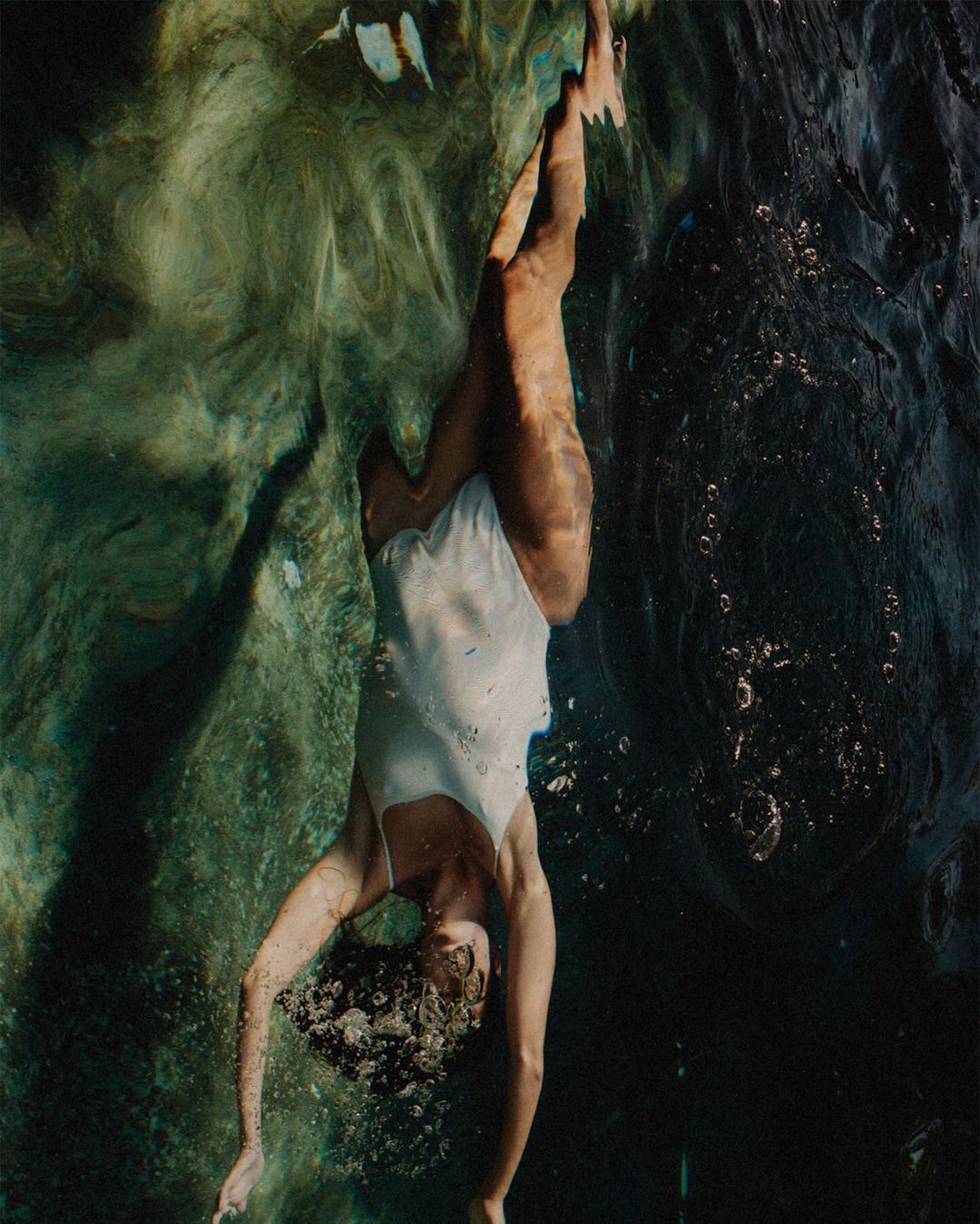 Underwater view of a woman in a white bathing suit, diving headfirst into dark, textured water with bright reflections and bubbles.