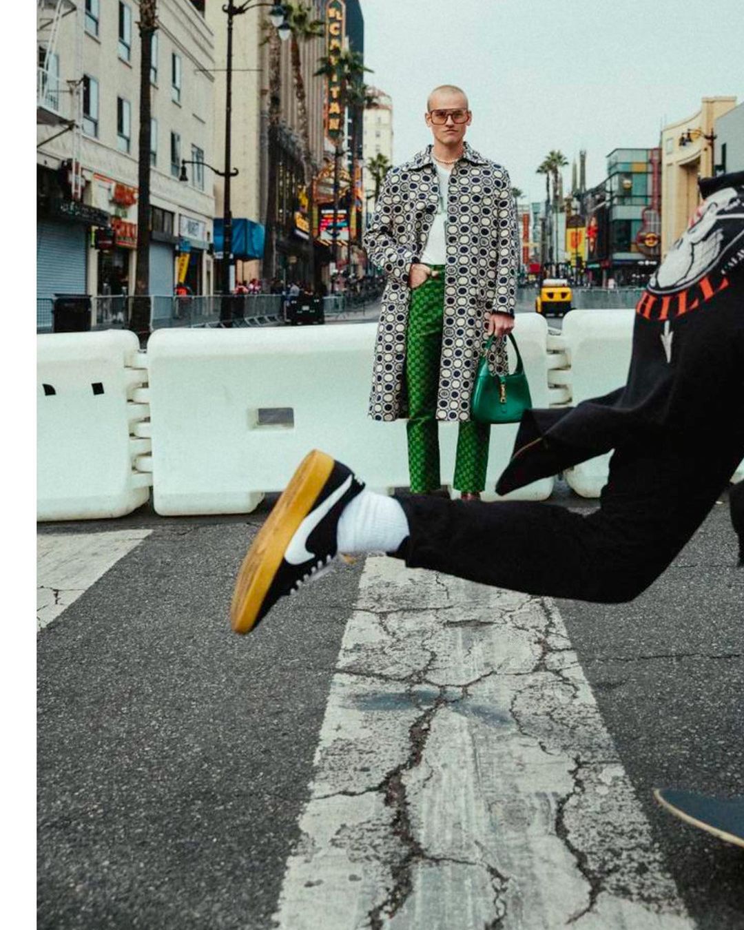 Fashionable man in patterned coat stands on a city street; a skateboarder moves in the foreground with a black and white sneaker.