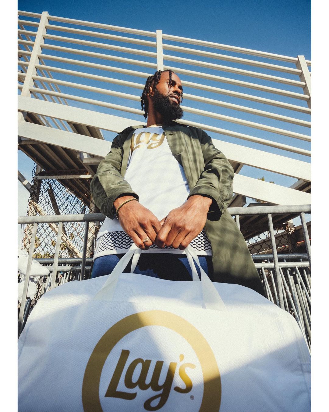 Adult male with a beard holding a large white Lay's potato chip bag near an outdoor sports bleacher with a clear blue sky overhead.