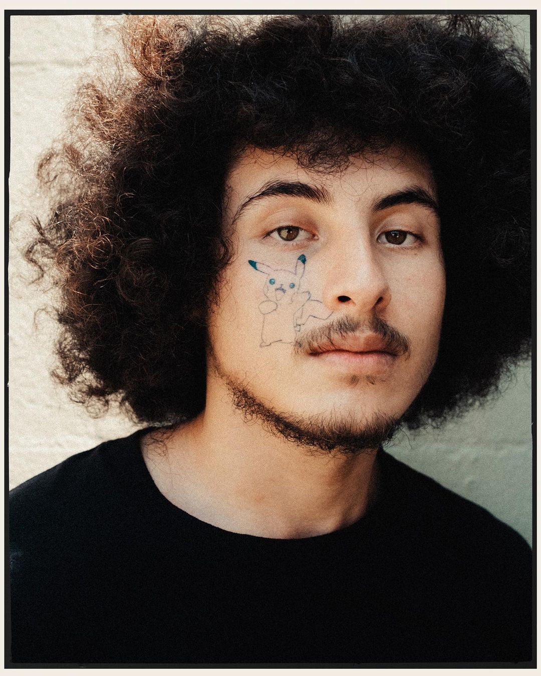 Close-up portrait of an adult man with an afro hairstyle and a Pikachu illustration on his face, wearing a black t-shirt against a textured wall.