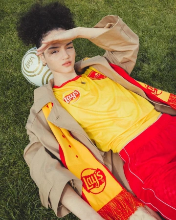 Woman in soccer jersey lies on grass with soccer ball. The jersey and soccer ball have Lay's potato chip logo. She wears a trench coat.