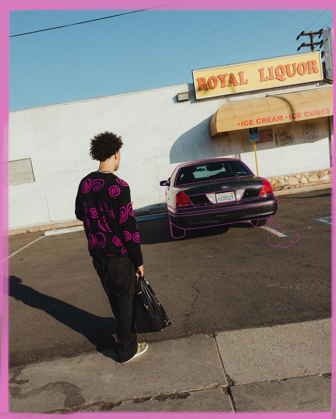Adult male stands near a parked car in an urban parking lot in front of a liquor store with a black shopping bag.