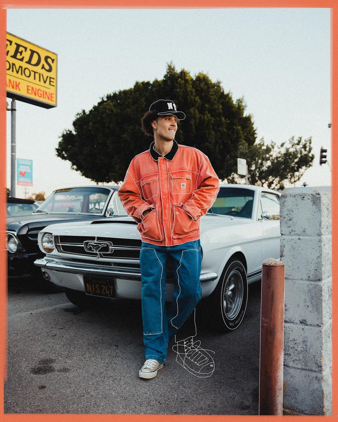 Young adult man in a coral jacket and jeans poses by a vintage white Ford Mustang car, with a Seeds Automotive sign in the background.