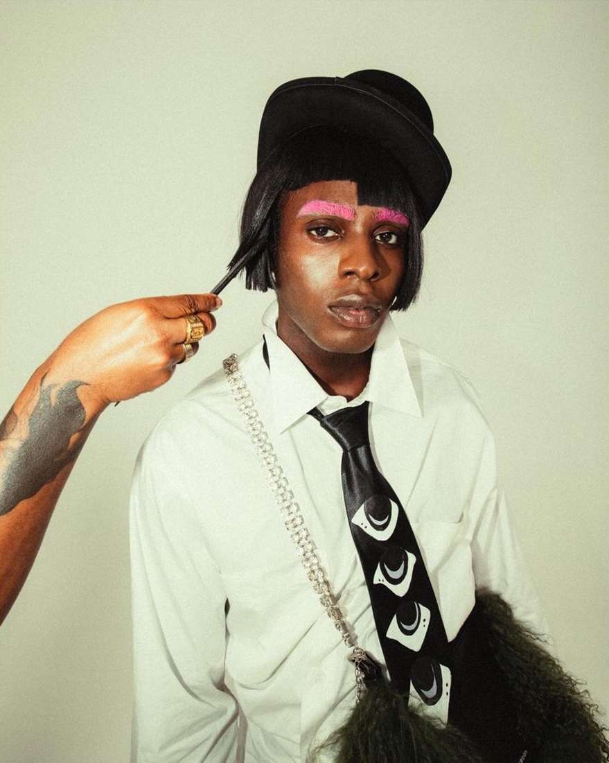 Person with pink eyebrows wearing a black hat and patterned tie, styled by a hand with gold rings, against a neutral background.
