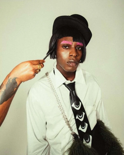 Person with pink eyebrows wearing a black hat and patterned tie, styled by a hand with gold rings, against a neutral background.
