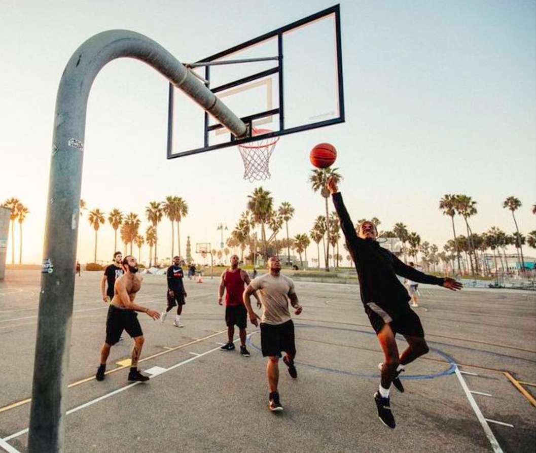 Action shot of a basketball game on an outdoor court with palm trees and a clear sky. Player is reaching for the ball near the hoop.