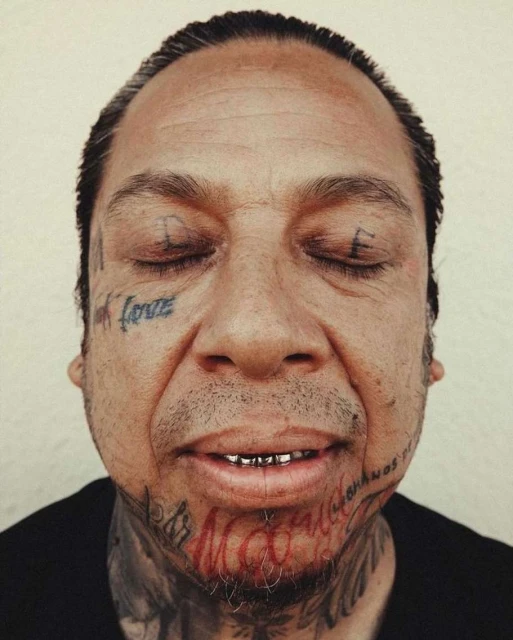 Portrait of an adult male with closed eyes, multiple face and neck tattoos, and gold teeth, set against a plain background.