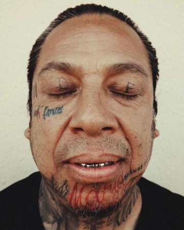 Portrait of an adult male with closed eyes, multiple face and neck tattoos, and gold teeth, set against a plain background.
