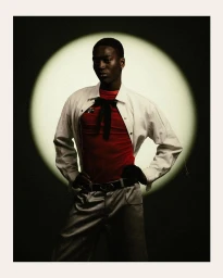 Portrait of a Black man standing against a circular spotlight backdrop, wearing a white jacket over a red shirt with a black bow.